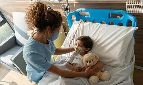 Boy lying down on hospital bed and sweet nurse comforting him both wearing facemasks during the COVID-19 pandemic