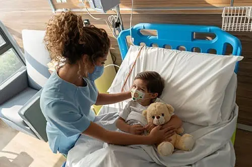 Boy lying down on hospital bed and sweet nurse comforting him both wearing facemasks during the COVID-19 pandemic