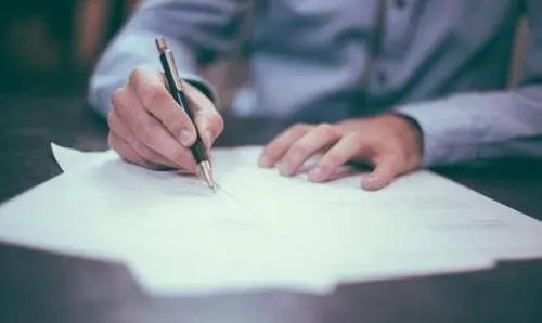 Person in a button up shift writing with a pen on a stack of papers