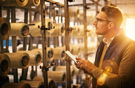 A man stands before rows of spools of thread on a industrial machine with a tablet in hand